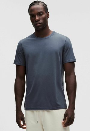 Uomo dalla pelle scura e capelli intrecciati che indossa una maglietta a maniche corte grigio scuro semplice e pantaloni chiari su uno sfondo semplice.