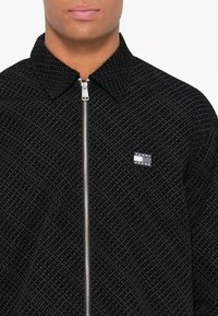 Juodas zip-up striukė, kuriai būdingas tekstūruotas raštas su prekės ženklu. Su apykakle ir maža prekės ženklo etikete kairėje pusėje.