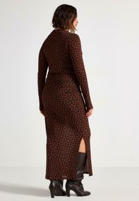 Lang, brun kjole med sorte prikker, der har lange ærmer og en sideslids. Blødt stof med en figursyet silhuet. Bæres med knæhøje støvler.