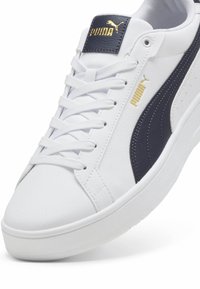 Hvide sneakers med en glat overflade, prydet med en marineblå stribe på siden og guldlogo i accenter. Klassisk snørebåndsdesign med en tyk sål.