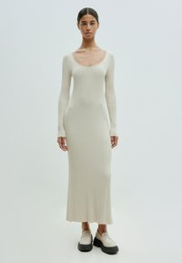 Crème ribgebreide jurk met lange mouwen en een ronde halslijn. Model draagt chunky loafers met zwarte zool. Eenvoudige silhouette, jurk tot op de vloer.