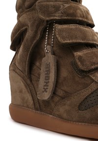 Καφέ σουέτ παπούτσι πλατφόρμα με λουράκια βέλκρο, διάτρητες λεπτομέρειες και μια ετικέτα από σουέτ σε μεταλλική αλυσίδα με μπίλια με την επιγραφή «BRONX».