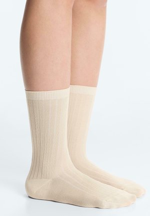 Jambes portant des chaussettes côtelées couleur crème, debout sur une surface blanche avec un fond blanc uni.