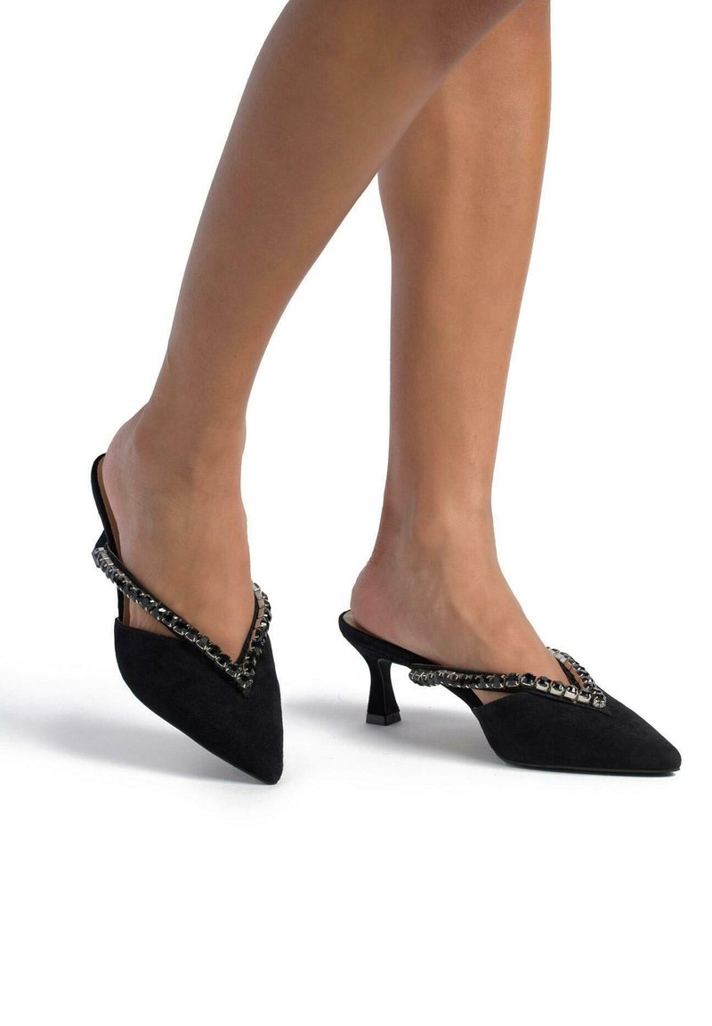 Le mule nere in suede presentano una punta affusolata e un design a forma di V, decorate con un accento di catena decorativa, poggianti su un tacco corto e solido.