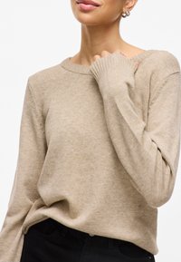 Pull en tricot beige, encolure ronde, poignets côtelés, texture douce, coupe décontractée, présenté avec une main reposant sur l'épaule, assorti à un pantalon noir.
