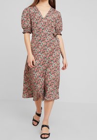 Žena v midi zavinovací šatech s krátkými puffovými rukávy a červeným a zeleným květinovým vzorem, spárovaných s černými sandály na otevřenou špičku.