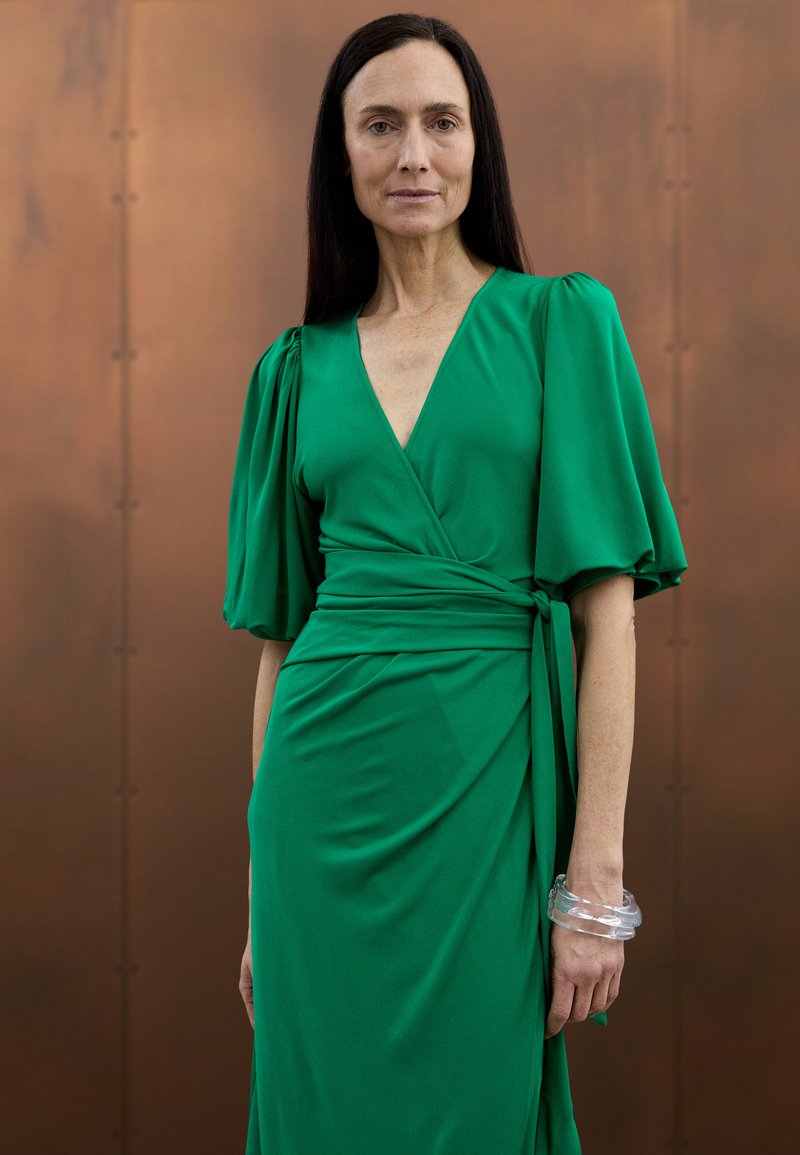 Green wrap dress with short puff sleeves and a deep V-neck. Features a tied waist and smooth texture. Accompanied by a clear bangle.