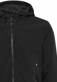 Chaqueta negra con capucha, cierre de cremallera frontal, tela texturizada, cordones ajustables y una pequeña etiqueta con logo en el lateral inferior. Forro interior suave.
