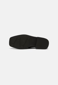 Semelle de chaussure noire à bout carré avec motif de bande de roulement, vue par le dessous sur un fond blanc uni.