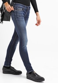 Vaqueros ajustados de mezclilla en color azul, con zonas desgastadas, diseño de cinco bolsillos y cintura de altura media. Combinados con zapatos negros y un bolso.