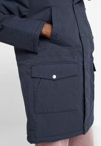Chaqueta acolchada de color navy con un bolsillo frontal con botón a presión, bolsillos laterales y una textura suave. El diseño incluye un cierre frontal con botones.