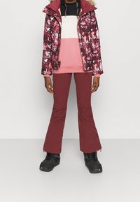 Kobieta w czerwonej, kwiatowej kurtce zimowej, różowym i beżowym swetrze z kolorowym blokiem, bordowych spodniach i czarnych butach stoi na jednolitym tle.