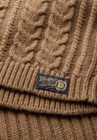 Berretto marrone lavorato a maglia con motivo a trecce, bordo a coste e un'etichetta rettangolare blu con testo e emblema dorati. Tessuto misto lana testurizzato.