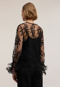 Blusa nera trasparente con ricamo floreale e dettagli perline. Presenta maniche arricciate e design posteriore con apertura a goccia. Tessuto liscio.