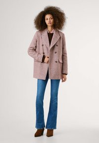 Manteau double boutonnage en chevron rose avec des boutons dorés, associé à un pull marron et un jean évasé bleu, sur fond neutre.