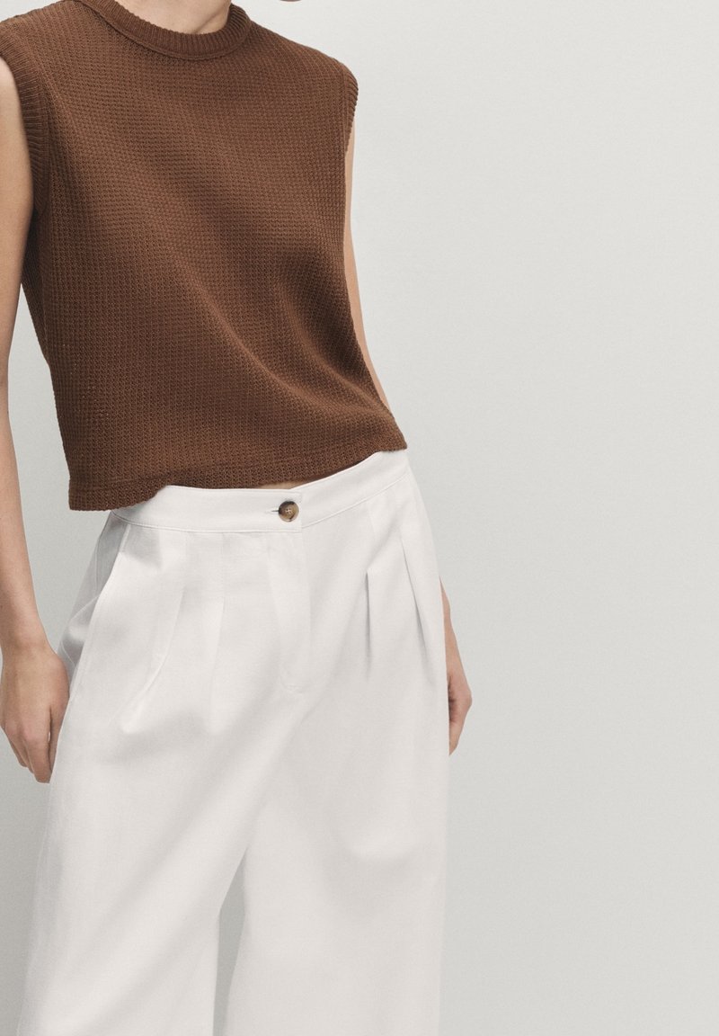 Bruine, mouwloze gebreide top met een gestructureerd oppervlak, gecombineerd met witte geplooide broek met een knoopdetail op de tailleband.