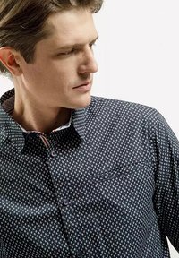 Ciemnoniebieska koszula z białym wzorem w kotwice, zapinana na guziki, z kołnierzykiem typu point i kieszenią na lewej stronie. Teksturowany materiał.