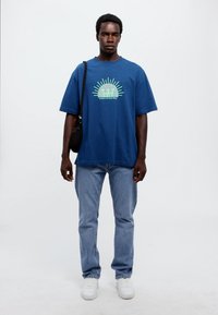Blå bomull t-skjorte med grønn grafisk trykk som viser en sol og tekst, kombinert med lyseblå denimjeans og hvite joggesko, svart skulderveske.