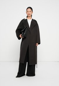 Abrigo largo de lana marrón oscuro con solapas grandes y cierre de botón único, combinado con pantalones anchos negros y un bolso de mano negro.