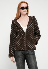 Brązowa kurtka z polaru z wzorzystą fakturą, kapturem i zamkiem błyskawicznym. Do tego czarne obcisłe jeansy i minimalistyczne okulary przeciwsłoneczne.