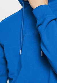 Primo piano di una persona che indossa un felpa blu brillante con testo in rilievo e cappucci metallici, mano che toca il mento.