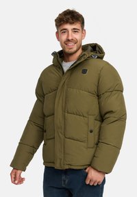 Mand smilende iført en olivengrøn pufferjakke med hætte og blå jeans, stående foran en hvid baggrund.
