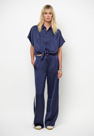 Vrouw die tegen een witte achtergrond staat, gekleed in een marineblauw satijnen shirt met korte mouwen en knopen, gestrikt in de taille, en bijpassende wijde broek.