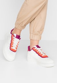 Sneakers bianche con accenti viola e arancione, caratterizzate da una tomaia in pelle liscia, punta forata e lacci piatti. Abbinati a pantaloni jogger beige.