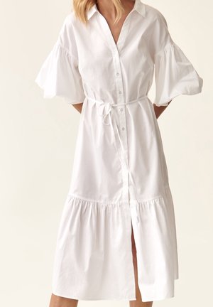 Vrouw draagt een wit midi-jurk met pofdrie-kwart mouwen, knopen aan de voorkant, een kraag en een ceintuur die is gestrikt in de taille.