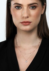 Collana a strati in oro rosa con un delicato pendente a forma di cuore adornato con strass, mostrata sul collo di un modello.