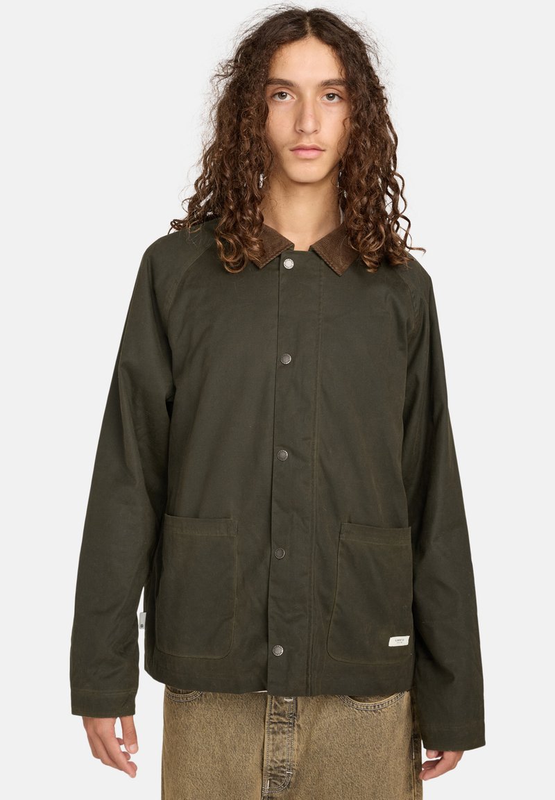 Junge Person mit langen lockigen Haaren, die eine dunkelgrüne Knopfjacket mit großen Fronttaschen und braunem Kragen sowie beigen Hosen trägt.