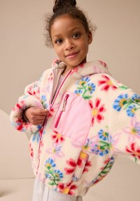 Giacca in pile color crema con motivi floreali colorati. Presenta una tasca con zip rosa, cappuccio e polsini elasticizzati per una vestibilità aderente.