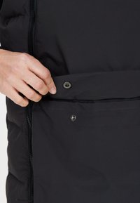 Sort polstret jakke med lynlås og trykknapper på frontlommen. Glat tekstur med en mat finish. Nærbillede af hånd, der betjener trykknappen.