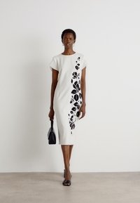 Abito bianco aderente con applicazioni floreali nere sul lato, maniche corte e design alla lunghezza del ginocchio; modella che tiene una piccola borsa nera.