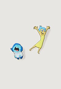 Dos pins de esmalte: un personaje con un suéter azul y una expresión triste, otro con un vestido amarillo y cabello azul, con los brazos levantados en señal de alegría.