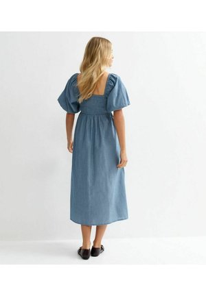 Femme aux longs cheveux blonds portant une robe midi bleue avec des manches bouffantes et un dos froncé, se tenant face à un mur blanc.