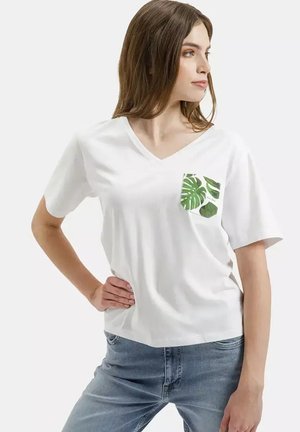 Młoda kobieta w białym T-shircie z dekoltem w serek i zieloną kieszenią z motywem tropikalnych liści, pozuje z jedną ręką na biodrze, w niebieskich dżinsach, patrząc na bok.