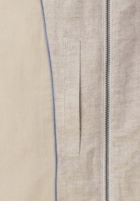 Gros plan sur une veste en tissu beige texturé avec une fermeture éclair verticale et une poche passepoilée assortie en tissu, accompagnée d'une doublure intérieure lisse de couleur tabac.