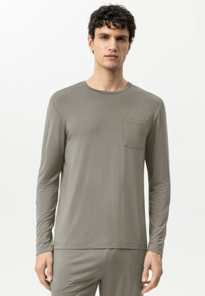 Mann mit lockigem dunklem Haar, der ein hellgraues Langarmhemd mit Brusttasche und passende Hose trägt, vor einem schlichten weißen Hintergrund stehend.