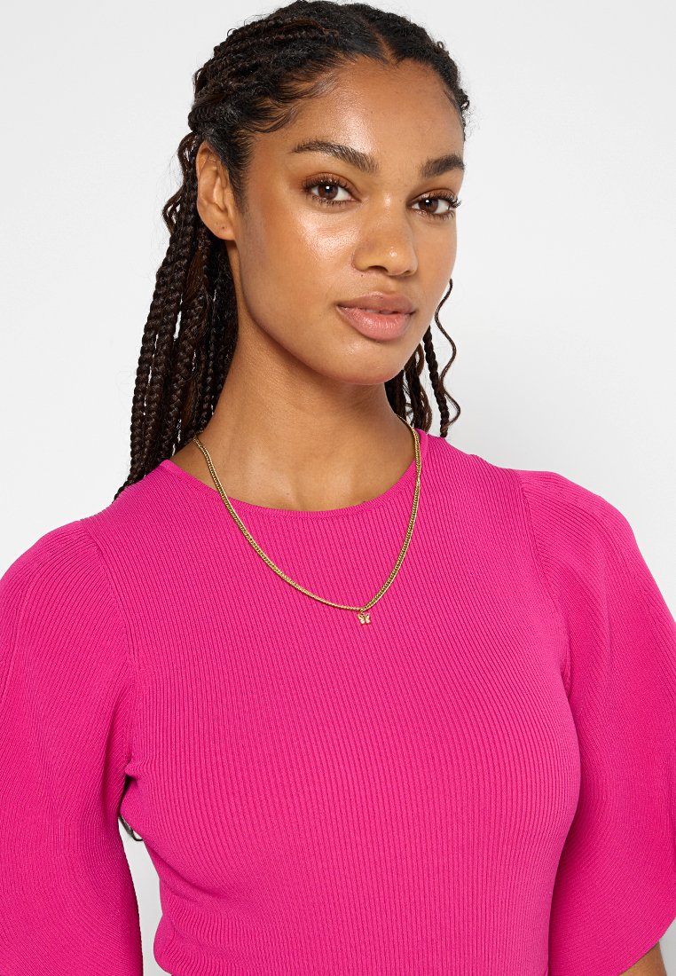 Rosa gerippter Pullover mit Puffärmeln, kombiniert mit einer zarten goldenen Kette, die einen kleinen Anhänger trägt. Hautfarbe ist mittel und die Haare sind geflochten.