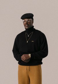 Maglione a collo alto nero con ricami dorati abbinato a pantaloni gialli con texture. Gli accessori includono occhiali e collane in oro a strati.