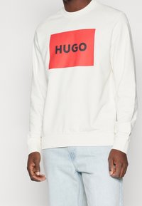 Felpa bianca con un grafico rettangolare rosso su cui è stampato "HUGO" in nero. Presenta polsini e fondo a costine e un design a girocollo.