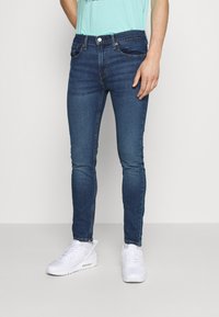 Jeans ajustados azul oscuro con desgaste descolorido y cinco bolsillos, combinados con una camisa turquesa y zapatillas blancas. Diseño sencillo, sin patrones visibles.