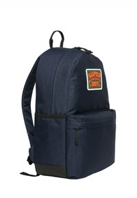 Sac à dos bleu marine en tissu avec une texture lisse. Il dispose d'une poche avant, d'accent noir et d'un patch blanc avec du texte rouge.