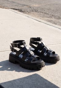 Ζευγάρι μαύρα chunky σανδάλια πλατφόρμας με λουράκια και αγκράφες τοποθετημένα σε ηλιόλουστο τσιμεντένιο πεζοδρόμιο κοντά σε ασφαλτοστρωμένο δρόμο.