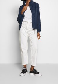 Chaqueta bomber azul marino, camiseta blanca, jeans rectos blancos y zapatillas de lona negras con suelas blancas; ajuste relajado con dobladillo deshilachado en los jeans.