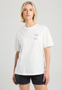 Valkoinen puuvillapaita lyhyillä hihoilla, jossa on violetti martini-lasi -grafiikka vasemmalla puolella. Yhdistetty tummiin shortseihin.