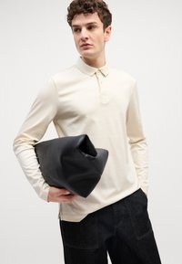 Bež polo majica dugih rukava s kragnom na gumbe, uparen s tamnoplavim trapericama, drži crnu kožnu torbicu. Glatka tekstura, minimalni dizajn.