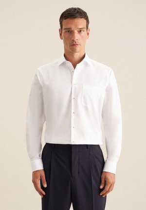 Witte button-up overhemd van gladde stof met een borstzakje en contrasterende knopen, gecombineerd met donkere, op maat gemaakte broek. Mouwen zijn lichtjes opgerold.