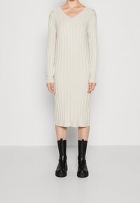 Vrouw in een crèmekleurige geribbelde gebreide midi-jurk met lange mouwen en zwarte chunky laarzen tot halverwege de kuit, staand tegen een eenvoudige muur.